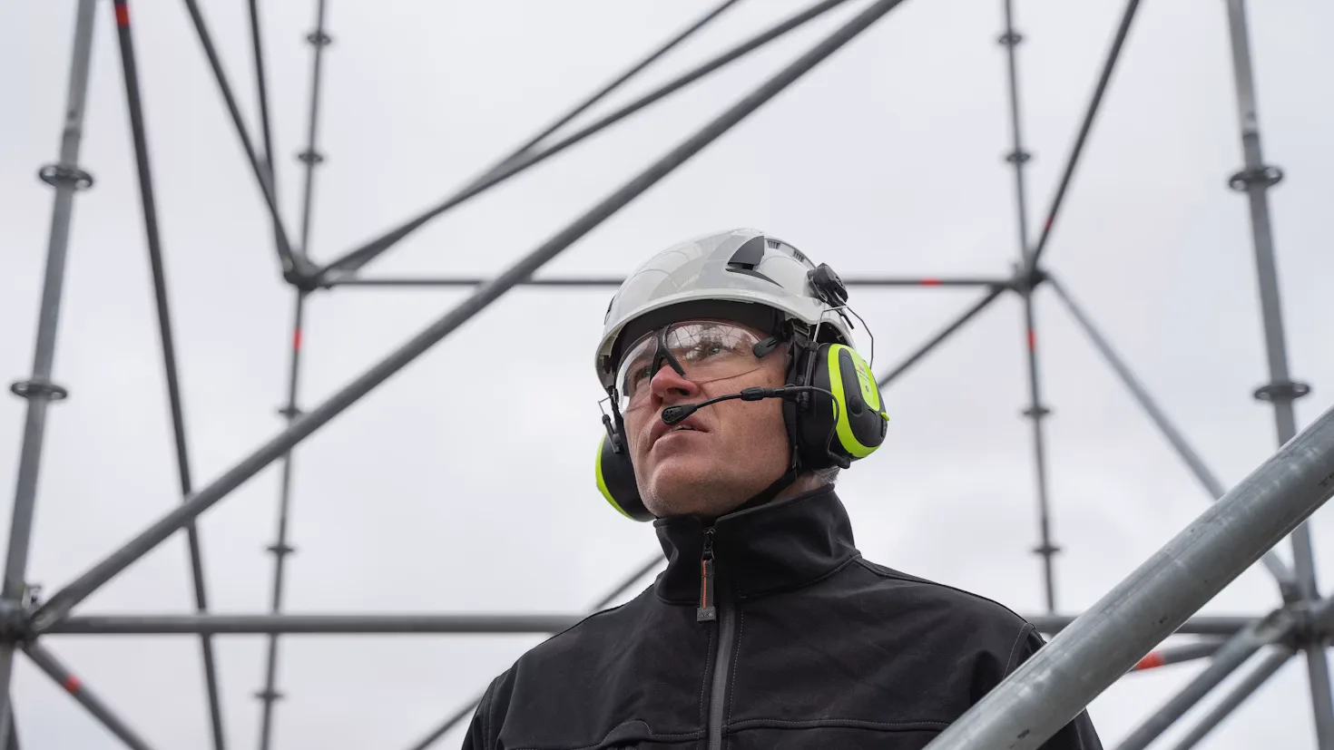 Un ouvrier du bâtiment casqué et avec un casque-micro, en pleine conversation devant un échafaudage.